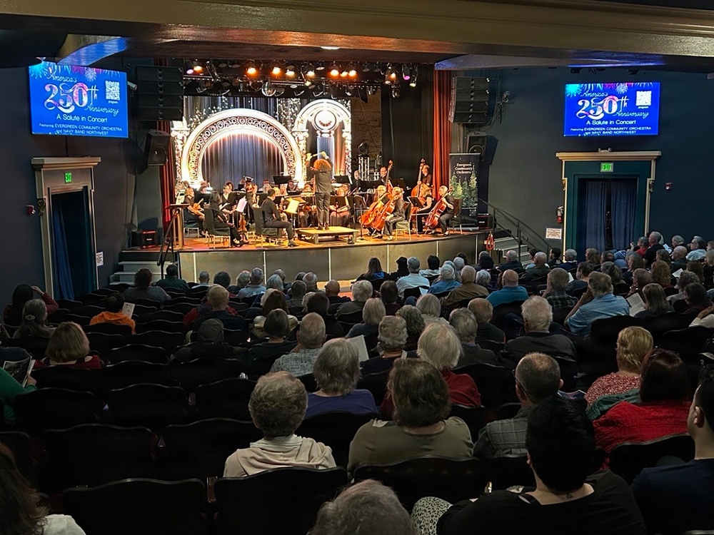 An orchestra concert is taking place in a mid-sized theater venue with a full audience seated in rows of dark chairs. Two digital display screens on either side of the stage show promotional graphics reading 'America's 250th Anniversary - A Salute in Concert' featuring the Evergreen Community Orchestra and US Navy Band Northwest. A conductor stands at the podium directing musicians on a decorated stage with an ornate arch backdrop.