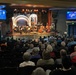 An orchestra concert is taking place in a mid-sized theater venue with a full audience seated in rows of dark chairs. Two digital display screens on either side of the stage show promotional graphics reading 'America's 250th Anniversary - A Salute in Concert' featuring the Evergreen Community Orchestra and US Navy Band Northwest. A conductor stands at the podium directing musicians on a decorated stage with an ornate arch backdrop.
