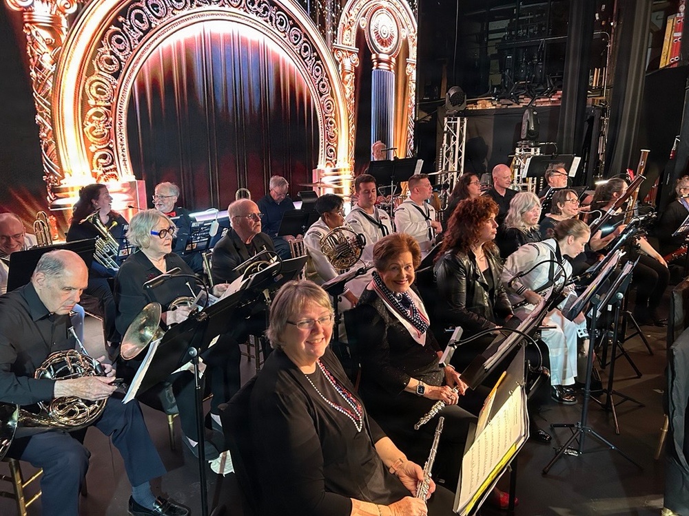 A large orchestra or concert band is assembled in what appears to be a theatrical venue, seated with music stands and instruments in front of an ornately decorated proscenium arch with red curtains and gold detailing. The musicians are predominantly dressed in black attire, with a few individuals in white naval-style uniforms visible in the middle rows. The setting suggests a pre-performance or rehearsal moment, with musicians holding or resting their instruments.