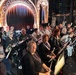 A large orchestra or concert band is assembled in what appears to be a theatrical venue, seated with music stands and instruments in front of an ornately decorated proscenium arch with red curtains and gold detailing. The musicians are predominantly dressed in black attire, with a few individuals in white naval-style uniforms visible in the middle rows. The setting suggests a pre-performance or rehearsal moment, with musicians holding or resting their instruments.