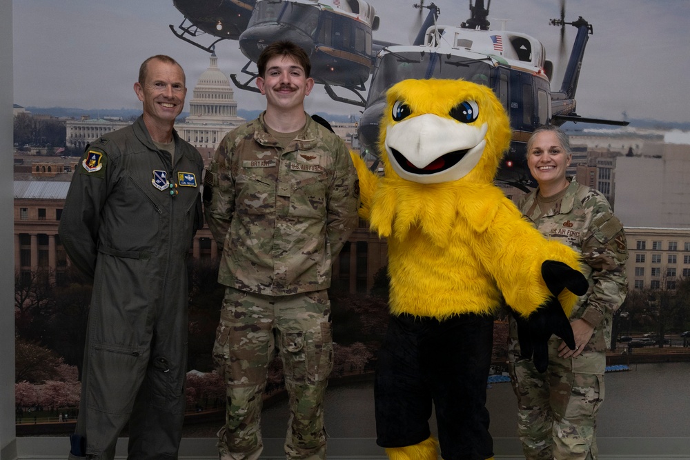 Three U.S. Air Force personnel pose for a photo with a large yellow bird mascot in front of a backdrop depicting military helicopters over Washington D.C. The group includes one person in an olive green flight suit with squadron patches, one in Army Combat Uniform (ACU/OCP) with a name tape reading 'BRYANT' and 'U.S. AIR FORCE', and one female service member also in OCP uniform. The setting appears to be an indoor military event or recruitment/public affairs function.