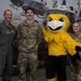 Three U.S. Air Force personnel pose for a photo with a large yellow bird mascot in front of a backdrop depicting military helicopters over Washington D.C. The group includes one person in an olive green flight suit with squadron patches, one in Army Combat Uniform (ACU/OCP) with a name tape reading 'BRYANT' and 'U.S. AIR FORCE', and one female service member also in OCP uniform. The setting appears to be an indoor military event or recruitment/public affairs function.