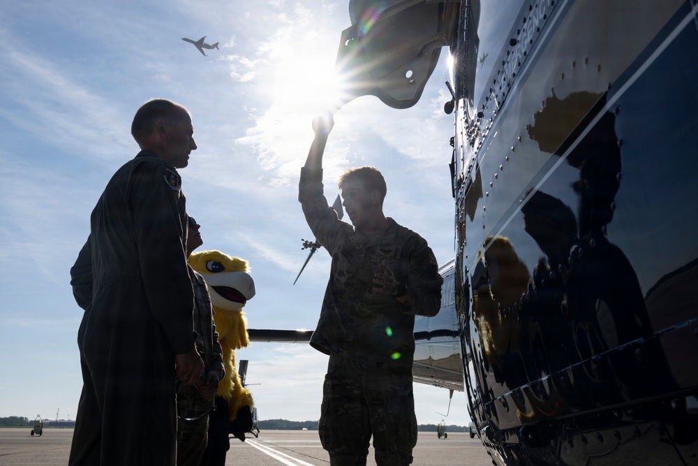 Military personnel are gathered on an airfield tarmac next to a large aircraft, with one service member pointing upward toward the aircraft's fuselage or wing. A child holding a yellow bird mascot/stuffed animal is present among the group. The scene is backlit by strong sunlight, creating silhouettes and lens flare effects.