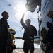 Military personnel are gathered on an airfield tarmac next to a large aircraft, with one service member pointing upward toward the aircraft's fuselage or wing. A child holding a yellow bird mascot/stuffed animal is present among the group. The scene is backlit by strong sunlight, creating silhouettes and lens flare effects.