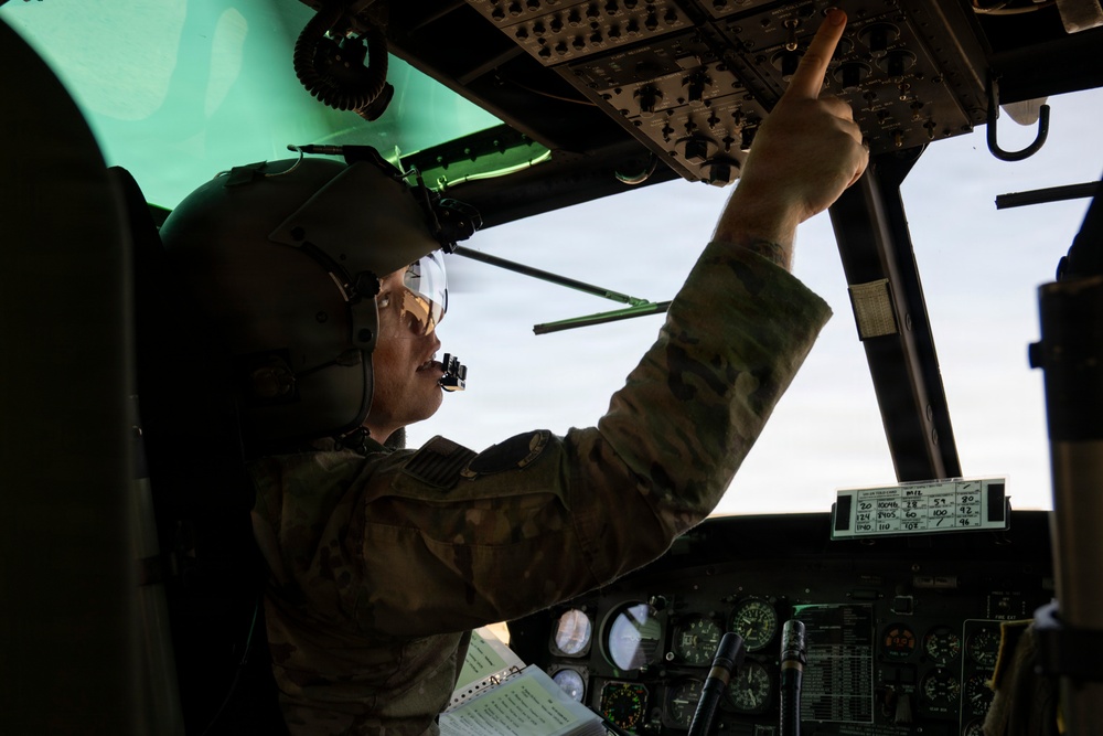 A military aviator in flight gear is seated in a helicopter cockpit, pointing upward at the overhead control panel. The individual is wearing a flight helmet with a boom microphone, clear visor, and camouflage uniform, suggesting a pre-flight or in-flight checklist procedure. The cockpit instrumentation and overhead switch panels are visible in the dimly lit interior.