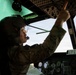 A military aviator in flight gear is seated in a helicopter cockpit, pointing upward at the overhead control panel. The individual is wearing a flight helmet with a boom microphone, clear visor, and camouflage uniform, suggesting a pre-flight or in-flight checklist procedure. The cockpit instrumentation and overhead switch panels are visible in the dimly lit interior.