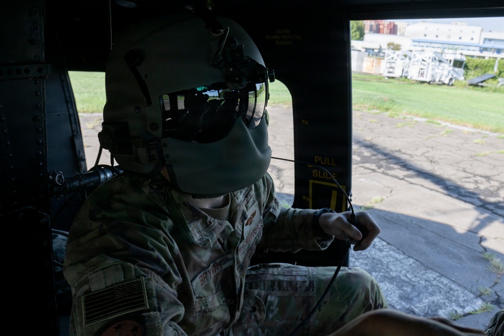 A U.S. military service member in full flight gear, including a helmet with visor and face mask, is seated inside what appears to be a military helicopter. The individual is wearing OCP (Operational Camouflage Pattern) uniform with a U.S. flag patch and a name tape reading 'BRYAN' visible on the chest. The aircraft door is open, revealing a ground-level view of a paved area, grass, and industrial structures in the background.