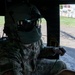A U.S. military service member in full flight gear, including a helmet with visor and face mask, is seated inside what appears to be a military helicopter. The individual is wearing OCP (Operational Camouflage Pattern) uniform with a U.S. flag patch and a name tape reading 'BRYAN' visible on the chest. The aircraft door is open, revealing a ground-level view of a paved area, grass, and industrial structures in the background.