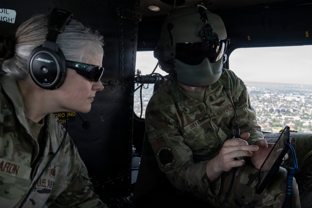 Two U.S. military personnel are seated inside a helicopter in flight, visible by the aerial cityscape through the window. The individual on the left wears U.S. Army uniform with Bose aviation headset and tactical sunglasses, while the individual on the right wears a U.S. Air Force uniform with a flight helmet and is operating a tablet device. The name tapes visible read 'ARON' (U.S. Army) and 'BRYAN' (U.S. Air Force), suggesting a joint-service operation.
