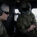 Two U.S. military personnel are seated inside a helicopter in flight, visible by the aerial cityscape through the window. The individual on the left wears U.S. Army uniform with Bose aviation headset and tactical sunglasses, while the individual on the right wears a U.S. Air Force uniform with a flight helmet and is operating a tablet device. The name tapes visible read 'ARON' (U.S. Army) and 'BRYAN' (U.S. Air Force), suggesting a joint-service operation.