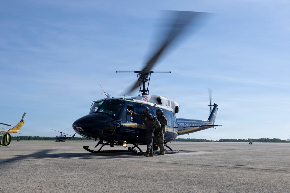 A dark blue and white 'United States of America' marked military helicopter, consistent with a UH-1 Huey variant, is on a tarmac with its rotors spinning. Two personnel in military flight gear and helmets are positioned near the open cockpit door, appearing to conduct pre-flight checks or a crew exchange. Additional aircraft and ground equipment are visible in the background.