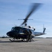 A dark blue and white 'United States of America' marked military helicopter, consistent with a UH-1 Huey variant, is on a tarmac with its rotors spinning. Two personnel in military flight gear and helmets are positioned near the open cockpit door, appearing to conduct pre-flight checks or a crew exchange. Additional aircraft and ground equipment are visible in the background.