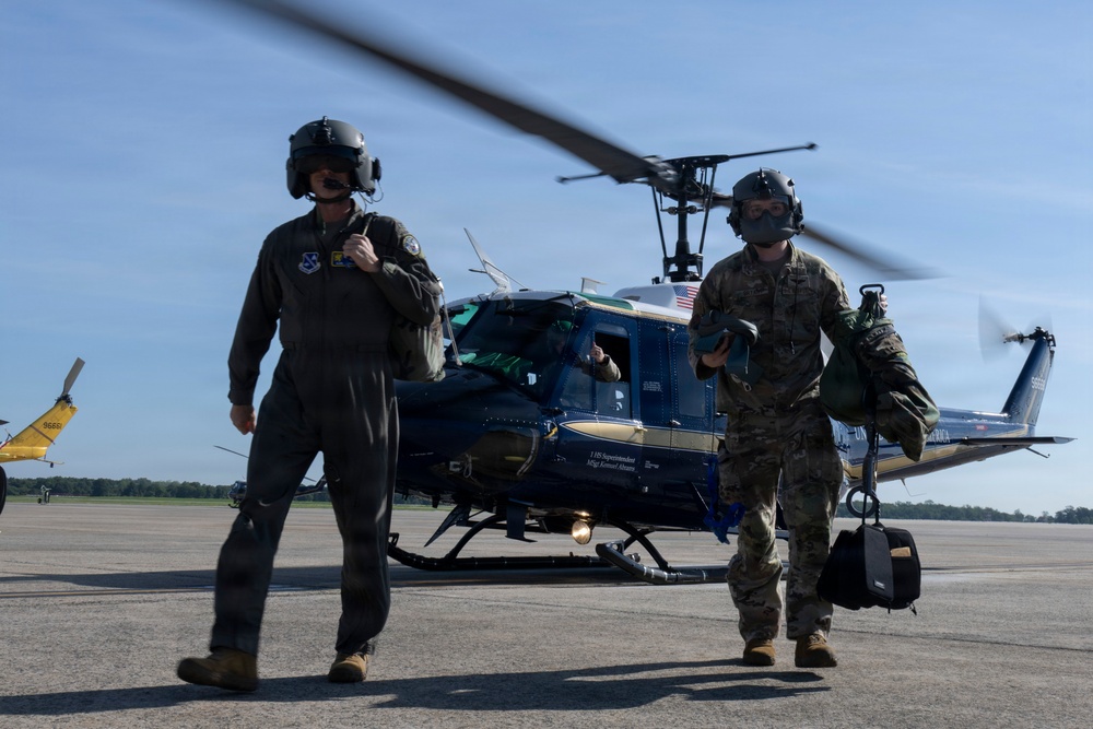 Two military personnel are walking away from a dark blue UH-1 Huey helicopter on a tarmac or airfield. The individual on the left is wearing a flight suit and helmet, while the person on the right is in camouflage uniform with a face mask and carrying equipment. A yellow aircraft is partially visible in the background on the left side of the frame.