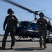 Two military personnel are walking away from a dark blue UH-1 Huey helicopter on a tarmac or airfield. The individual on the left is wearing a flight suit and helmet, while the person on the right is in camouflage uniform with a face mask and carrying equipment. A yellow aircraft is partially visible in the background on the left side of the frame.