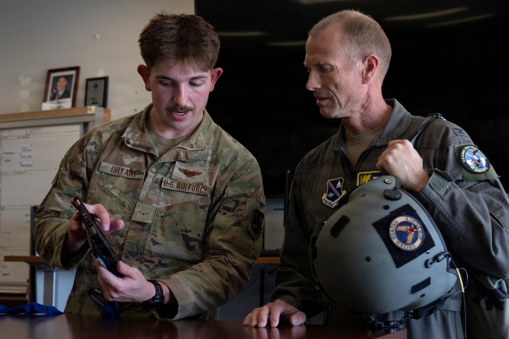 Two U.S. Air Force personnel are reviewing content on a tablet device at a desk or table. The individual on the left wears an OCP (Operational Camouflage Pattern) uniform with a name tape reading 'BRYANT' and U.S. Air Force insignia, while the individual on the right wears an olive flight suit with multiple patches and is holding a military flight helmet. They appear to be engaged in a briefing or equipment review session.
