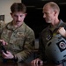 Two U.S. Air Force personnel are reviewing content on a tablet device at a desk or table. The individual on the left wears an OCP (Operational Camouflage Pattern) uniform with a name tape reading 'BRYANT' and U.S. Air Force insignia, while the individual on the right wears an olive flight suit with multiple patches and is holding a military flight helmet. They appear to be engaged in a briefing or equipment review session.