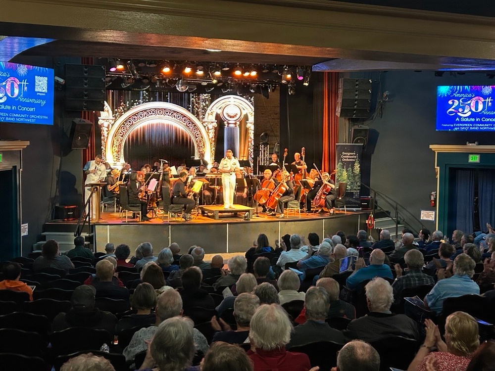 An orchestra concert is taking place in a decorated theater venue, featuring the Evergreen Community Orchestra and Navy Band Northwest in a performance titled 'America's 250th Anniversary - A Salute in Concert.' A person in a white naval uniform stands at center stage, likely serving as a speaker or conductor, while musicians with string and wind instruments are seated on stage. The theater is filled with a large audience, predominantly older adults, watching the performance.