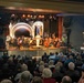 An orchestra concert is taking place in a decorated theater venue, featuring the Evergreen Community Orchestra and Navy Band Northwest in a performance titled 'America's 250th Anniversary - A Salute in Concert.' A person in a white naval uniform stands at center stage, likely serving as a speaker or conductor, while musicians with string and wind instruments are seated on stage. The theater is filled with a large audience, predominantly older adults, watching the performance.