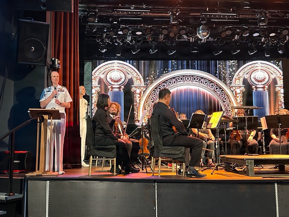 A military officer in white dress uniform speaks at a podium on a theater stage while an orchestra is seated behind them. The setting appears to be a formal concert or ceremony held in a performance venue with elaborate stage lighting and a decorative LED backdrop displaying ornate arches. A second uniformed individual stands nearby while string musicians hold their instruments at rest.