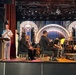 A military officer in white dress uniform speaks at a podium on a theater stage while an orchestra is seated behind them. The setting appears to be a formal concert or ceremony held in a performance venue with elaborate stage lighting and a decorative LED backdrop displaying ornate arches. A second uniformed individual stands nearby while string musicians hold their instruments at rest.