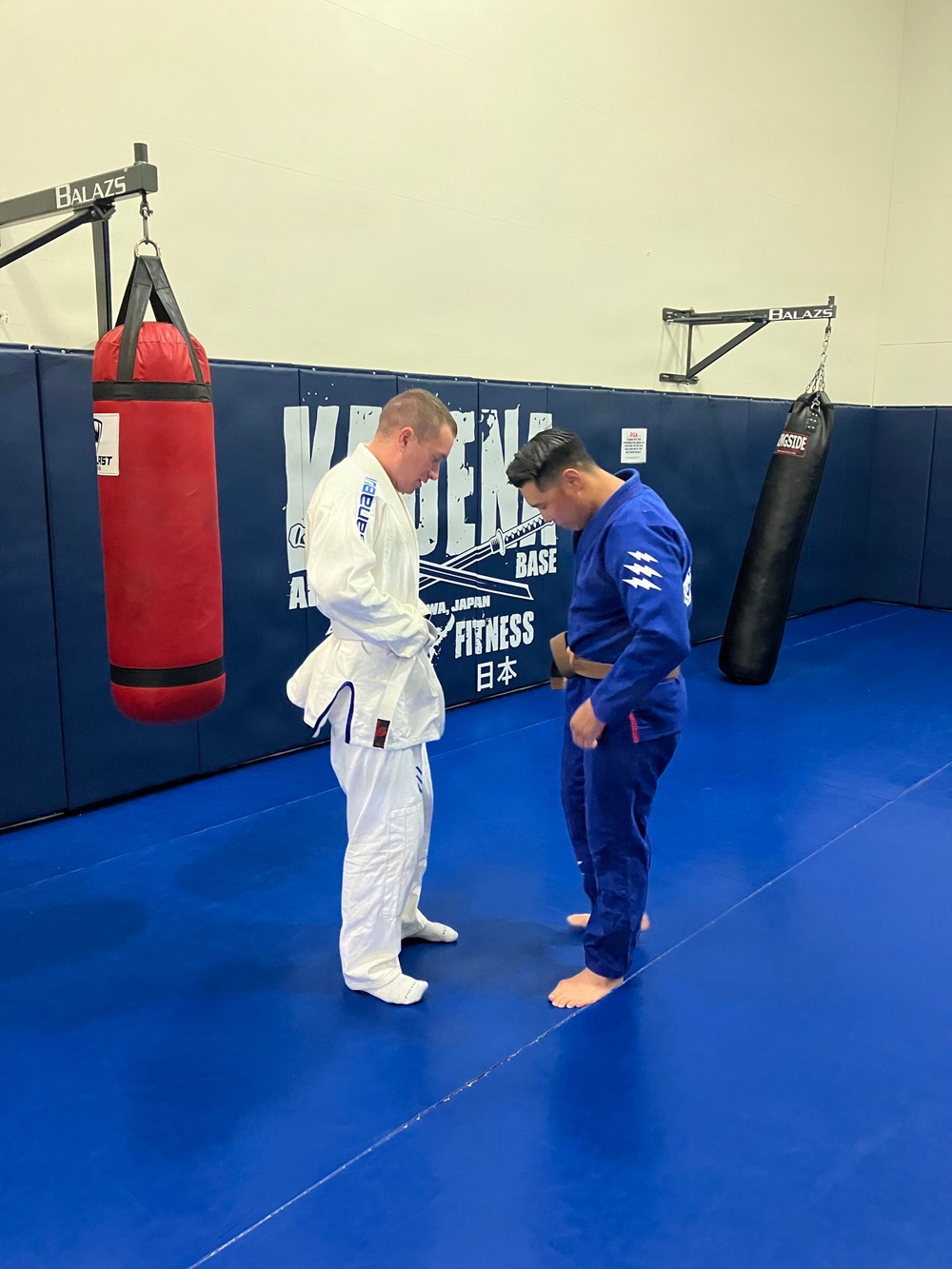 Two practitioners in Brazilian Jiu-Jitsu gis are bowing to each other on a blue mat in a martial arts gym. One wears a white gi with a blue belt, and the other wears a blue gi with a brown belt. The setting appears to be a military base fitness facility in Okinawa, Japan.
