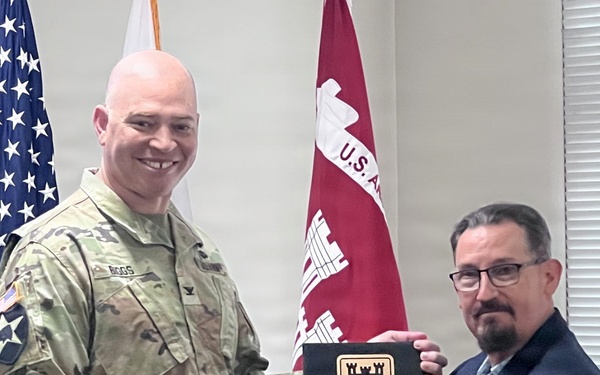 A U.S. Army soldier in camouflage uniform and a civilian in a dark suit are shaking hands while jointly holding a 'Gold Castle Award' plaque presented to the Japan Engineer District. The ceremony takes place in front of an American flag and a U.S. Army Corps of Engineers flag. The award appears to recognize completion of safety or occupational health management system actions.