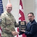 A U.S. Army soldier in camouflage uniform and a civilian in a dark suit are shaking hands while jointly holding a 'Gold Castle Award' plaque presented to the Japan Engineer District. The ceremony takes place in front of an American flag and a U.S. Army Corps of Engineers flag. The award appears to recognize completion of safety or occupational health management system actions.