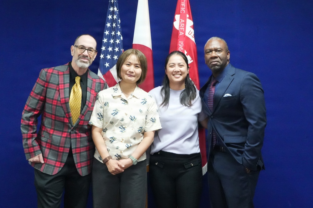 Four individuals are posing together for a formal group photograph in front of a blue backdrop with flags. The flags visible include the United States flag, the Japanese flag, and what appears to be a U.S. Army Pacific Area command flag. The setting suggests an official or ceremonial occasion.