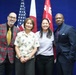 Four individuals are posing together for a formal group photograph in front of a blue backdrop with flags. The flags visible include the United States flag, the Japanese flag, and what appears to be a U.S. Army Pacific Area command flag. The setting suggests an official or ceremonial occasion.