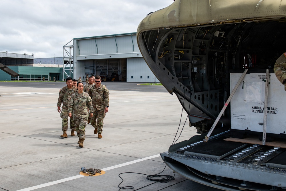 A group of military personnel in camouflage uniforms are walking on an airfield tarmac toward a large military aircraft with its cargo ramp open. The scene takes place at what appears to be a military airfield with hangars visible in the background. A white cargo container labeled 'HANDLE WITH CARE - REUSABLE CONTAINER' is visible loaded in the aircraft.