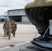 A group of military personnel in camouflage uniforms are walking on an airfield tarmac toward a large military aircraft with its cargo ramp open. The scene takes place at what appears to be a military airfield with hangars visible in the background. A white cargo container labeled 'HANDLE WITH CARE - REUSABLE CONTAINER' is visible loaded in the aircraft.