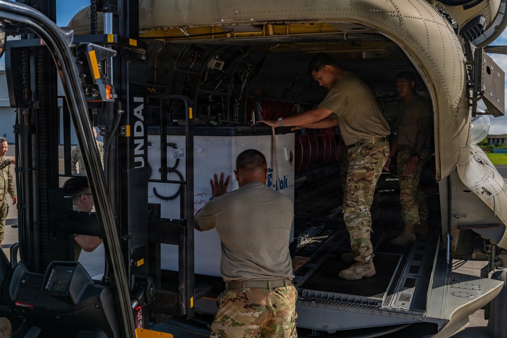 Military personnel in camouflage uniforms are loading cargo into what appears to be a large military aircraft. A Hyundai forklift is being used to position a large white container or pallet into the aircraft's cargo bay. Multiple service members are coordinating the loading operation on an airfield tarmac.