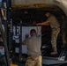 Military personnel in camouflage uniforms are loading cargo into what appears to be a large military aircraft. A Hyundai forklift is being used to position a large white container or pallet into the aircraft's cargo bay. Multiple service members are coordinating the loading operation on an airfield tarmac.