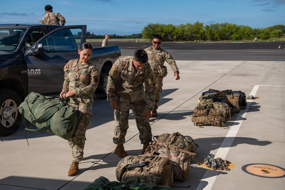 A group of military personnel in MultiCam/OCP camouflage uniforms are handling tactical gear and rucksacks on what appears to be an airfield tarmac. Several large military rucksacks and equipment bags are laid out on the ground, with soldiers actively moving and inspecting them. A Ram 1500 pickup truck is parked nearby, and the setting appears to be a warm, tropical or subtropical location based on the vegetation in the background.