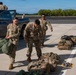 A group of military personnel in MultiCam/OCP camouflage uniforms are handling tactical gear and rucksacks on what appears to be an airfield tarmac. Several large military rucksacks and equipment bags are laid out on the ground, with soldiers actively moving and inspecting them. A Ram 1500 pickup truck is parked nearby, and the setting appears to be a warm, tropical or subtropical location based on the vegetation in the background.