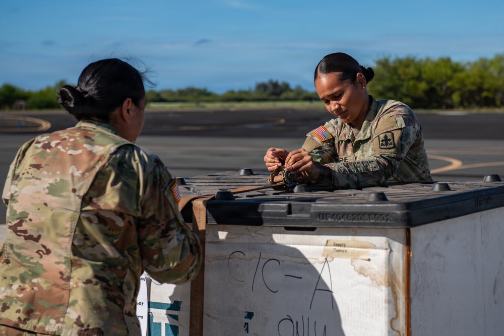 Two female U.S. Army soldiers in OCP (Operational Camouflage Pattern) uniforms are working together at an outdoor location, likely an airfield or tarmac. They appear to be handling or securing equipment on or near a large military shipping container or pallet. The setting features a clear blue sky, green treeline, and paved surface consistent with a military airfield.
