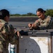 Two female U.S. Army soldiers in OCP (Operational Camouflage Pattern) uniforms are working together at an outdoor location, likely an airfield or tarmac. They appear to be handling or securing equipment on or near a large military shipping container or pallet. The setting features a clear blue sky, green treeline, and paved surface consistent with a military airfield.