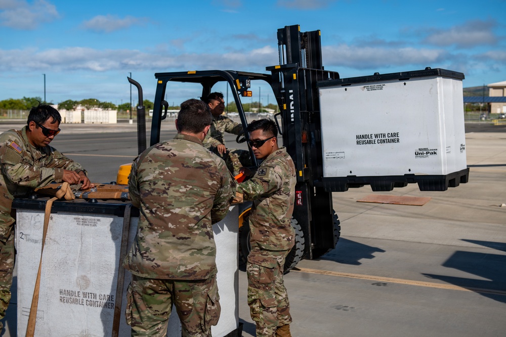 Military personnel in MultiCam/OCP camouflage uniforms are handling reusable shipping containers on what appears to be a military airfield or logistics area. A Hyundai forklift is being used to move a white container labeled 'Handle With Care Reusable Container' and branded 'Uni-Pak'. Four soldiers are visible, with one operating the forklift and three others managing containers on the ground.