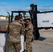 Military personnel in MultiCam/OCP camouflage uniforms are handling reusable shipping containers on what appears to be a military airfield or logistics area. A Hyundai forklift is being used to move a white container labeled 'Handle With Care Reusable Container' and branded 'Uni-Pak'. Four soldiers are visible, with one operating the forklift and three others managing containers on the ground.