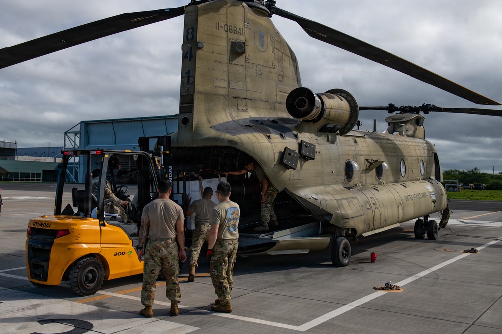 A United States Army CH-47 Chinook helicopter (tail number 11-08841) is being loaded or unloaded on a tarmac using a Hyundai 30D-9 forklift. Several soldiers in MultiCam/OCP uniforms are supervising and assisting the cargo operation at what appears to be a military airfield. The overcast sky and tropical vegetation in the background suggest a Pacific or tropical location.