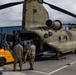 A United States Army CH-47 Chinook helicopter (tail number 11-08841) is being loaded or unloaded on a tarmac using a Hyundai 30D-9 forklift. Several soldiers in MultiCam/OCP uniforms are supervising and assisting the cargo operation at what appears to be a military airfield. The overcast sky and tropical vegetation in the background suggest a Pacific or tropical location.
