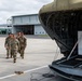 A group of military personnel in camouflage uniforms are walking on an airfield tarmac toward a large military transport aircraft with its rear cargo ramp open. A white cargo container labeled 'HANDLE WITH CARE - REUSABLE CONTAINER' is visible inside the aircraft's cargo bay. The scene takes place near a military hangar facility under overcast skies.