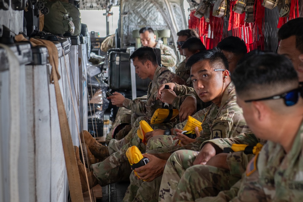 A group of military personnel in camouflage uniforms are seated inside what appears to be a military transport aircraft. The soldiers are holding yellow objects that appear to be safety or emergency equipment, possibly life vests or flotation devices. The interior of the aircraft shows typical military cargo configuration with equipment secured along the walls.