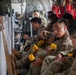 A group of military personnel in camouflage uniforms are seated inside what appears to be a military transport aircraft. The soldiers are holding yellow objects that appear to be safety or emergency equipment, possibly life vests or flotation devices. The interior of the aircraft shows typical military cargo configuration with equipment secured along the walls.