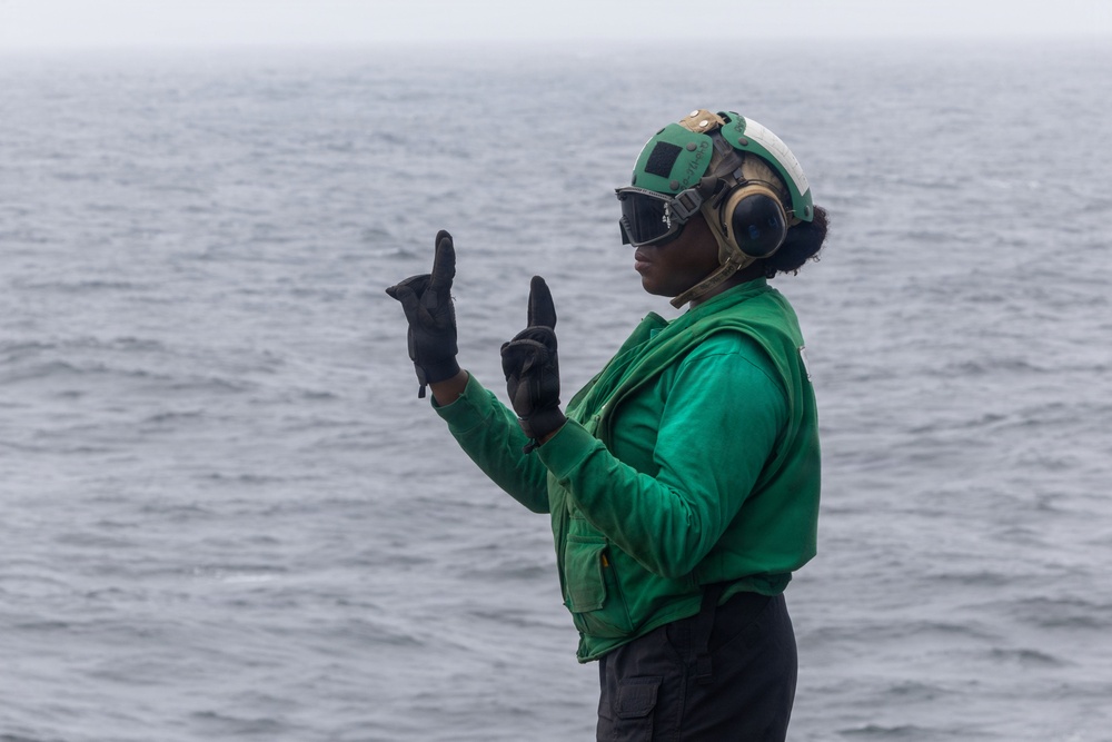 A naval flight deck crew member wearing a green jersey stands on what appears to be an aircraft carrier flight deck with the ocean visible in the background. The individual is using hand signals, a standard communication method used in high-noise flight deck environments. The green jersey indicates a role associated with aircraft handling, catapult, or arresting gear crews.