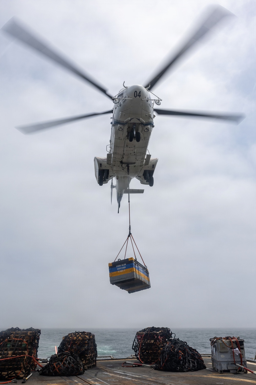 A military helicopter numbered '04' hovers over a ship's deck while conducting a vertical replenishment (VERTREP) operation, lowering a cargo container suspended by sling load. The container appears to be marked 'USNS Alan Shepard' in blue and yellow lettering. Multiple netted cargo pallets are visible on the ship's deck with the ocean visible in the background.
