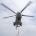 A military helicopter numbered '04' hovers over a ship's deck while conducting a vertical replenishment (VERTREP) operation, lowering a cargo container suspended by sling load. The container appears to be marked 'USNS Alan Shepard' in blue and yellow lettering. Multiple netted cargo pallets are visible on the ship's deck with the ocean visible in the background.