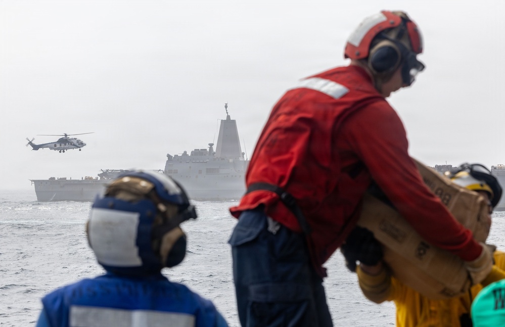 Naval personnel in colored safety gear observe a replenishment-at-sea (RAS) or vertical replenishment (VERTREP) operation from the deck of a ship. A military helicopter is airborne between two vessels on open, overcast seas. Crew members are handling cargo boxes as part of the logistics transfer operation.