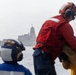 Naval personnel in colored safety gear observe a replenishment-at-sea (RAS) or vertical replenishment (VERTREP) operation from the deck of a ship. A military helicopter is airborne between two vessels on open, overcast seas. Crew members are handling cargo boxes as part of the logistics transfer operation.