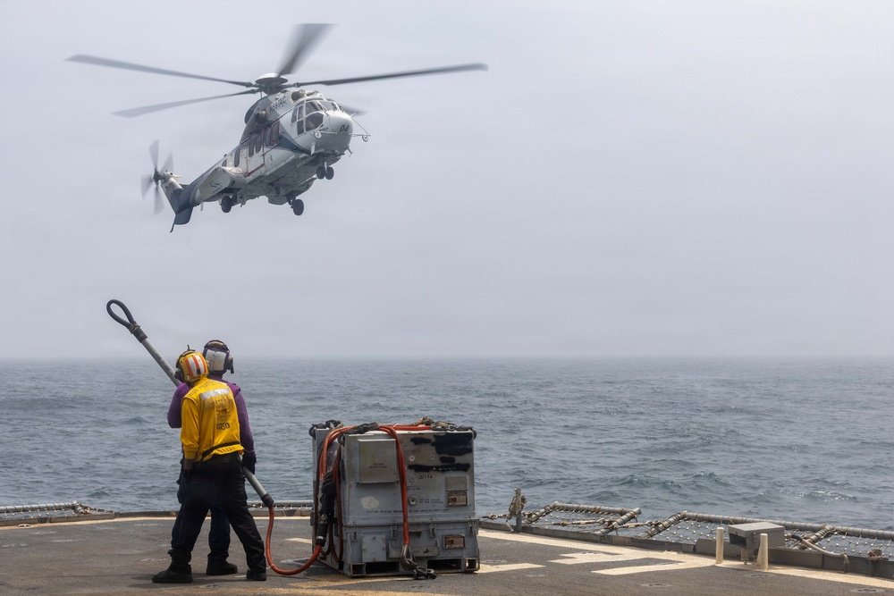 A military helicopter is hovering above the flight deck of a naval vessel at sea, while two deck crew members in safety gear appear to be preparing for a fueling or landing operation. One crew member in a yellow vest is holding what appears to be a grounding wand or fuel hose toward the helicopter. A metal equipment unit with orange hoses is positioned on the deck nearby.